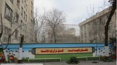 حیاط مدرسه پوشیده از درختان کهنسال با نیمکت‌های سبز و دیوار نقاشی‌شده به متن فارسی، محیط آرام و سبز برای فعالیت‌های در فضای باز دانش‌آموزان