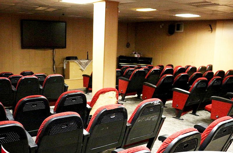 سالن مراسم و رویدادهای مدرسه با صندلی‌های قرمز و سیاه و پرده نمایش