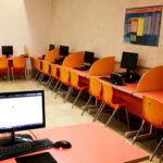 اتاق کامپیوتر و فناوری اطلاعات مدرسه با میز‌های قرمز، صندلی‌های نارنجی و رایانه‌های رومیزی