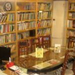 کتابخانه مدرسه با قفسه‌های بلند پُر از کتاب‌ها، میز و صندلی‌های مطالعه، و رایانه